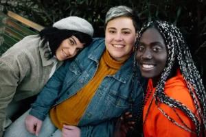 Three friends smile warmly while sitting close together. One wears a beret, another a denim jacket and mustard sweater, and the third a bright orange hoodie.