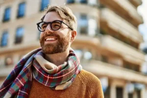 A smiling man with a beard and glasses, wearing a plaid scarf and orange sweater, stands outdoors in a city. The scene is bright and cheerful.