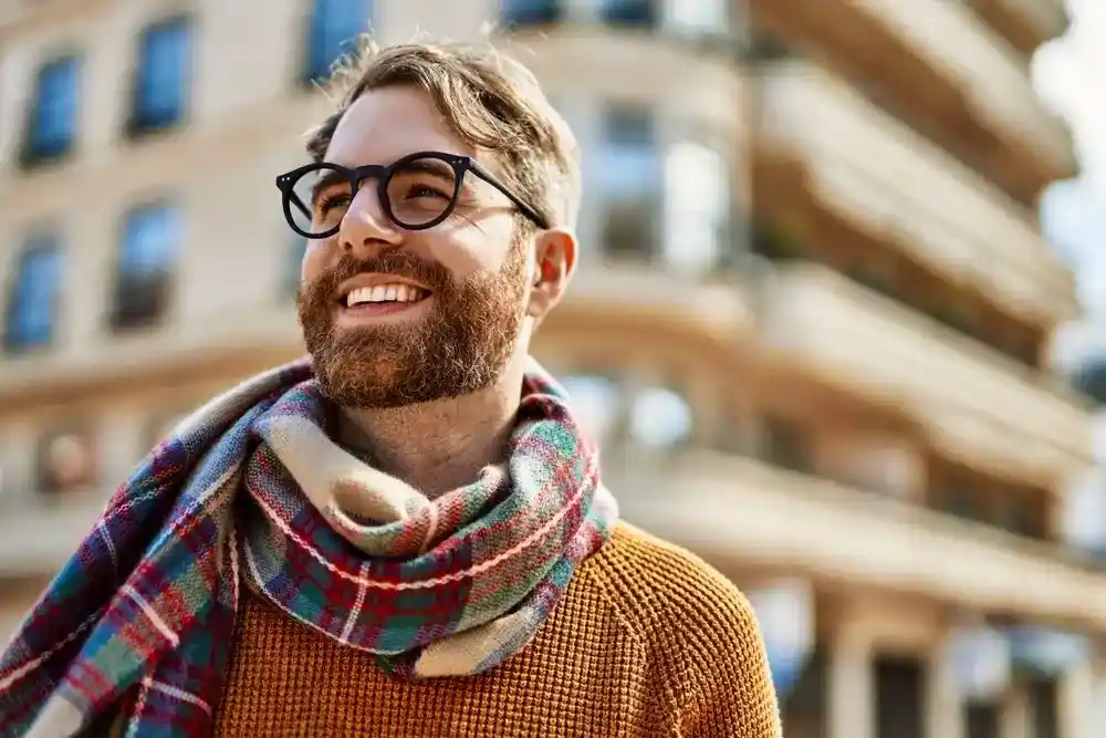 A smiling man with a beard and glasses, wearing a plaid scarf and orange sweater, stands outdoors in a city. The scene is bright and cheerful.
