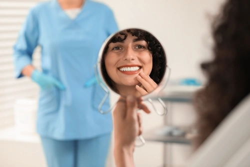 A woman smiles while holding a mirror, reflecting her face and pointing at her teeth. A dental professional in blue scrubs and gloves stands behind her.