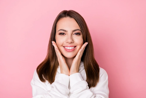 Smiling woman with long brown hair, wearing a white sweater, gently holds her face with both hands. She stands against a pink background, conveying a cheerful mood.