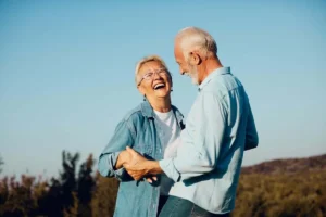 Elderly couple joyfully dancing outdoors, holding hands and laughing under a clear blue sky, conveying happiness and togetherness.