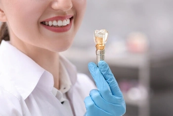 A smiling woman in a white coat holds a dental implant model with blue-gloved hand, conveying professionalism and dental care expertise.