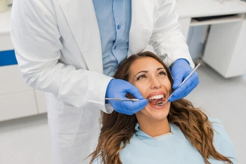 A dentist wearing blue gloves examines a smiling woman’s teeth with dental tools in a clinic, conveying a sense of care and professionalism.