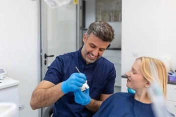 A dentist in a blue uniform and gloves smiles while showing a dental mold to a patient. The patient, wearing a protective bib, looks on attentively.