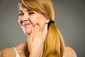 A woman with long blonde hair in a ponytail holds her jaw, grimacing in pain, against a neutral background, suggesting discomfort from a toothache.