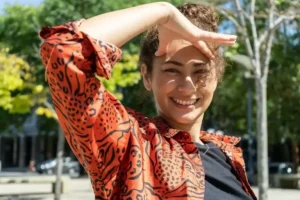 Smiling woman with curly hair wearing a vibrant orange patterned shirt holds her hand above her eyes for shade, standing outdoors on a sunny day.