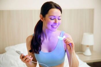A woman in a light blue tank top smiles while using a teeth whitening device emitting a blue light. She is in a cozy bedroom setting with soft lighting.