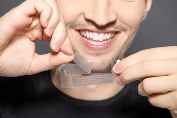 A person smiles broadly, showing white teeth while applying a transparent whitening strip on their upper teeth, suggesting a focus on dental care and hygiene.