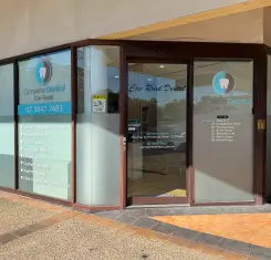 Street view of a dental clinic entrance with glass doors and windows. Signage displays clinic name and contact information, creating a welcoming atmosphere.