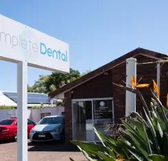 A small brick dental clinic with a "Complete Dental" sign, surrounded by parked cars and tropical plants, under a clear blue sky.