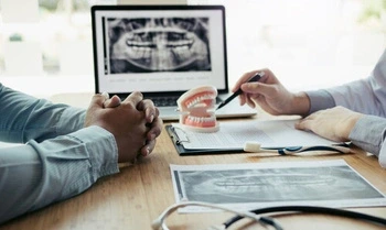 Two people at a dentist's office discuss dental X-rays. A dental model and clipboard are on the table, with a laptop displaying an X-ray in the background.
