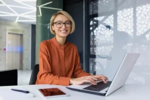 Smiling individual with glasses and short blonde hair, wearing an orange blouse, typing on a laptop in a modern office with geometric lighting.