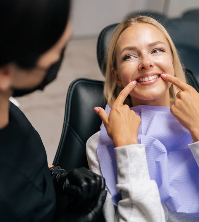 A woman smiles, pointing at her teeth while sitting in a dentist's chair. She wears a blue dental bib, and a person in black gloves watches her.