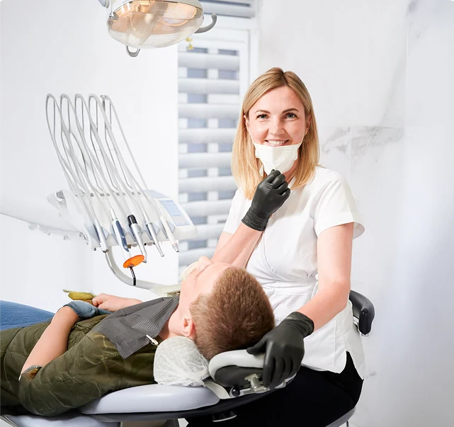 A smiling dentist with gloves and a mask is seated beside a reclining patient in an examination chair.