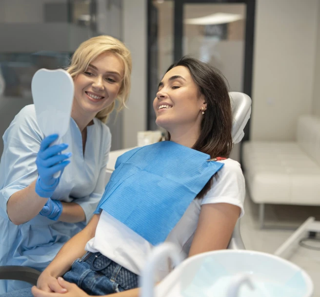 A dentist in blue gloves shows a female patient her smile in a hand mirror. Both are smiling, creating a relaxed and happy atmosphere in the dental office.