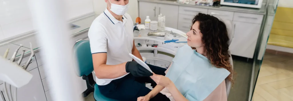A dentist in a mask and gloves consults with a patient, holding a tablet. The patient, wearing a bib, listens attentively in a modern dental office.