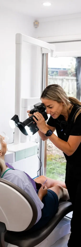 A dental professional photographs an elderly patient in a clinic. The patient is seated, and the setting is well-lit