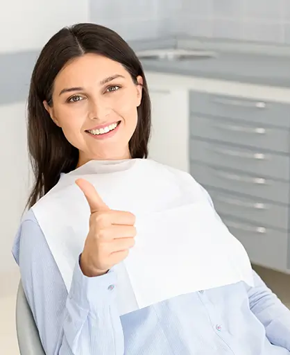 A woman in a dental office gives a thumbs-up, smiling warmly. She wears a dental bib, expressing confidence and satisfaction with her experience.