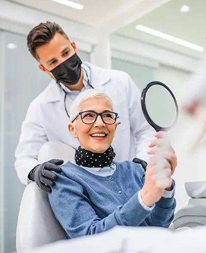 A smiling woman with short gray hair and glasses looks in a handheld mirror at the dentist's office.