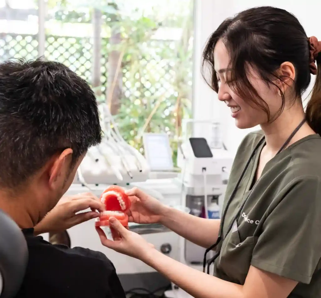 A dental professional in scrubs smiles while showing a model of teeth to a seated patient.