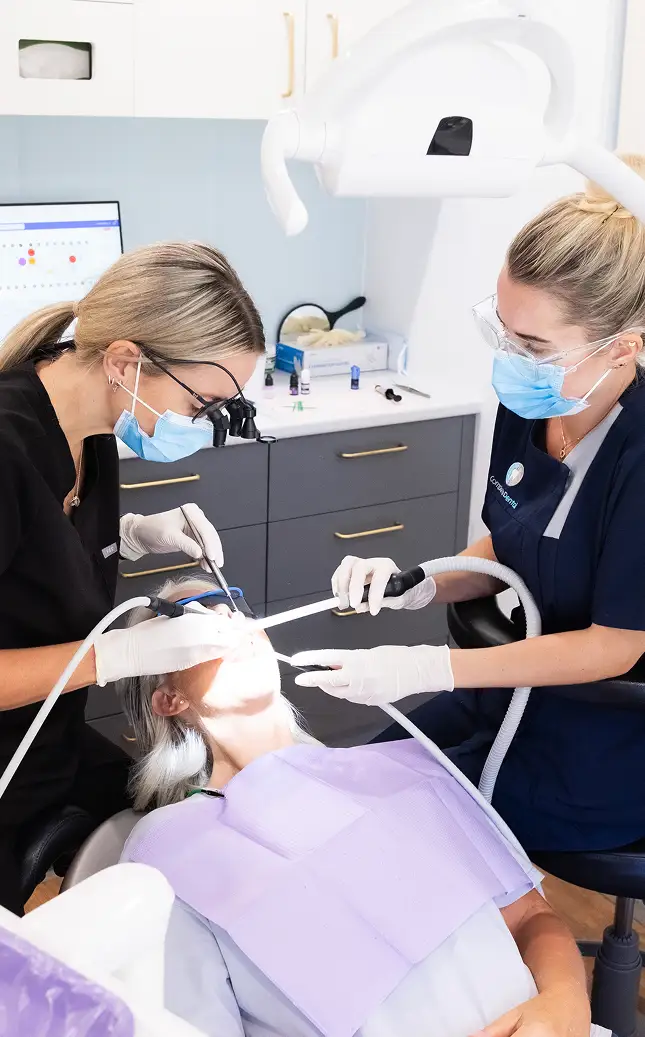 Two dental professionals wearing masks and gloves attend to a patient in a clinic.