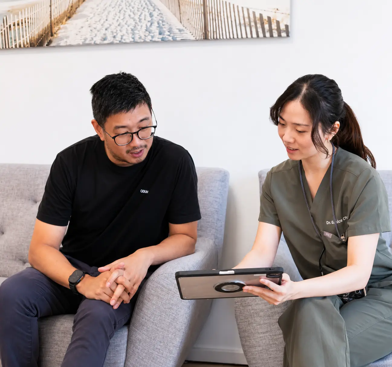 A woman in scrubs shows a tablet to a seated man in a black shirt on a gray couch.