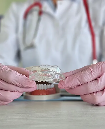 A dentist in a white coat and pink gloves holds a clear dental aligner above a model of teeth, conveying professionalism and dental care focus.