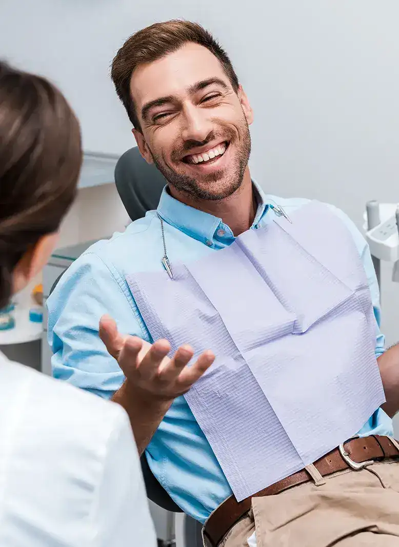 A man in a dentist's chair, wearing a bib, smiles and gestures with an open hand.