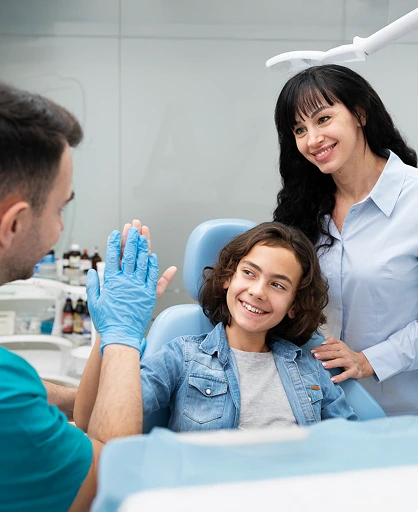 A dentist and a woman smile at a child sitting in a dental chair. The dentist high-fives the child, creating a friendly and reassuring atmosphere.