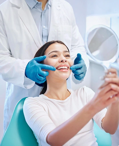 A smiling woman sits in a dentist's chair holding a mirror, as a dentist wearing blue gloves gently examines her teeth, creating a positive, reassuring atmosphere.
