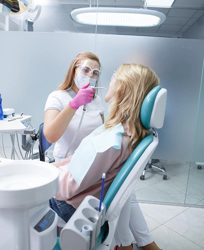 Dentist wearing gloves and a face shield attends to a patient in a modern dental clinic. The atmosphere is clinical and professional.