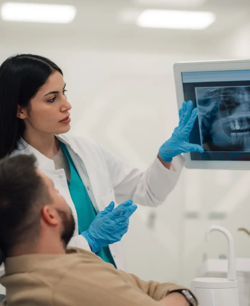 A dentist in a white coat and blue gloves shows a dental X-ray to a seated patient in a bright clinic, explaining the image with a focused expression.