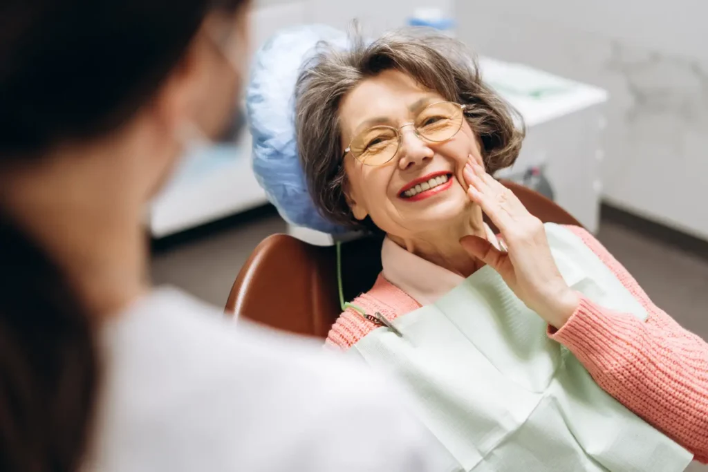 An elderly woman in glasses smiles warmly while sitting in a dental chair. She touches her cheek with satisfaction. A dentist is visible in soft focus.