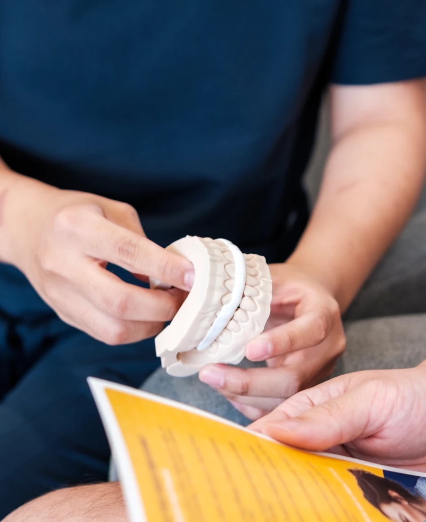 A person holds a dental model, pointing at the teeth, with another person holding a yellow pamphlet nearby.