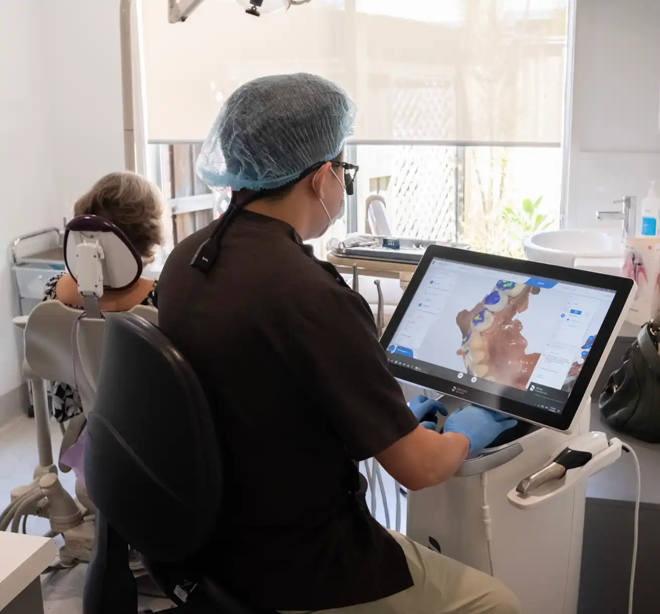 A dentist in a mask and cap uses a digital scanner in a modern clinic. A patient sits in a dental chair, creating a focused and professional atmosphere.