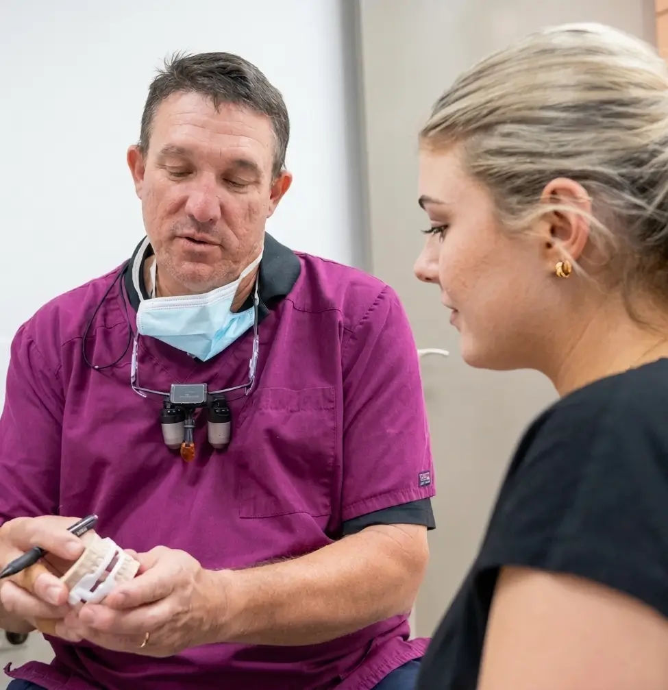 Dentist in a maroon uniform explains a dental model to a patient in a black shirt. Both are focused.
