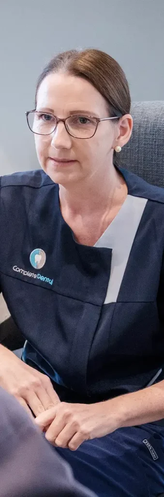 A woman in glasses and a navy dental uniform sits attentively, hands folded.