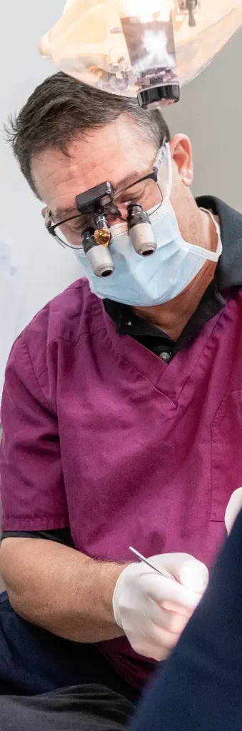 A dentist in maroon scrubs, wearing magnifying glasses and a mask.
