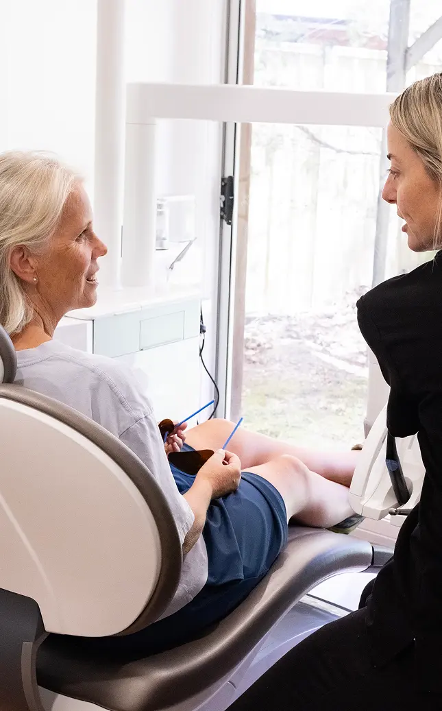 A woman with white hair sits relaxed in a dental chair, smiling, while a blonde dentist converses with her beside a large window
