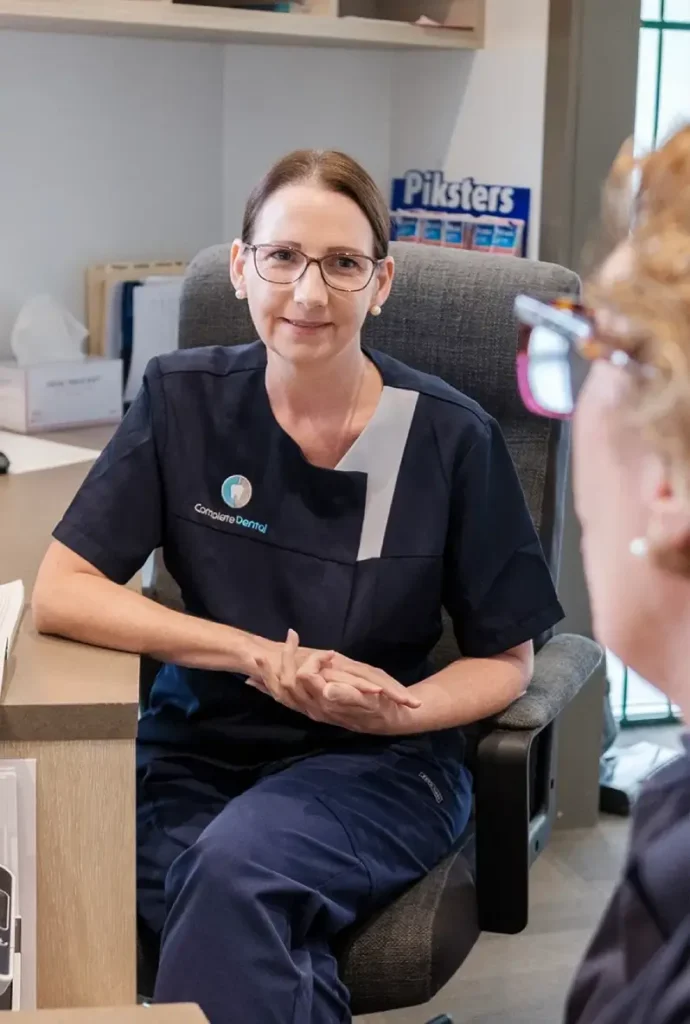A dental professional in a navy uniform sits at a desk, attentively listening to someone out of frame. The setting is calm and professional.
