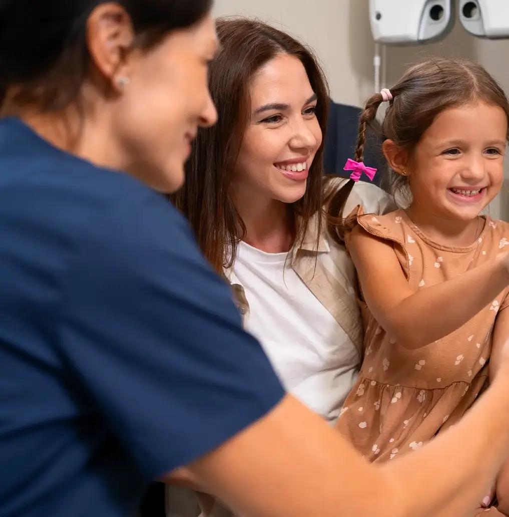 A smiling mother and daughter sit together in a medical office. The young girl, wearing a pink bow, is being examined by a nurse in blue scrubs.