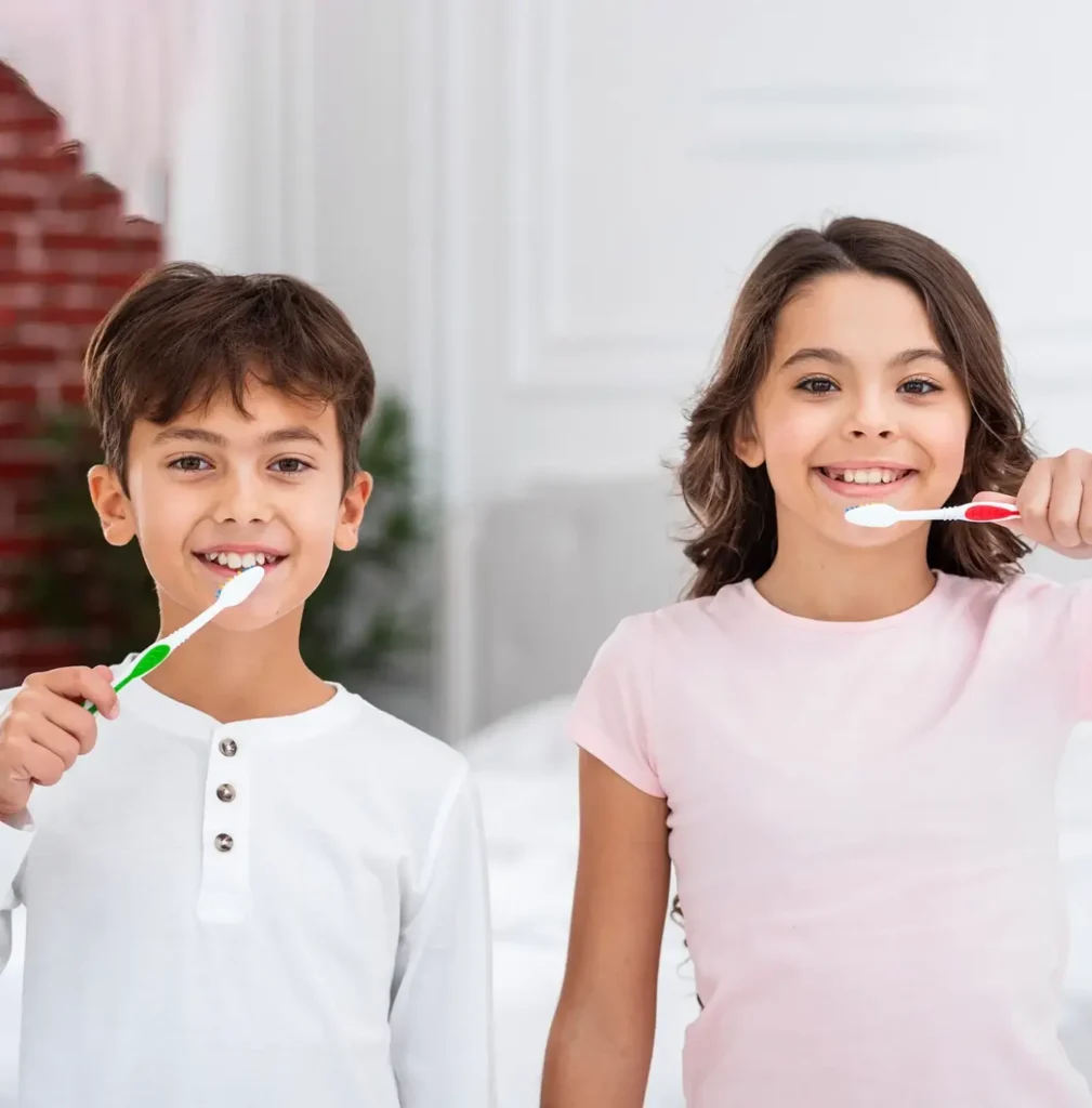 A young boy and girl are smiling while brushing their teeth with colorful toothbrushes. They stand in a bright, cozy room with a relaxed, joyful expression.