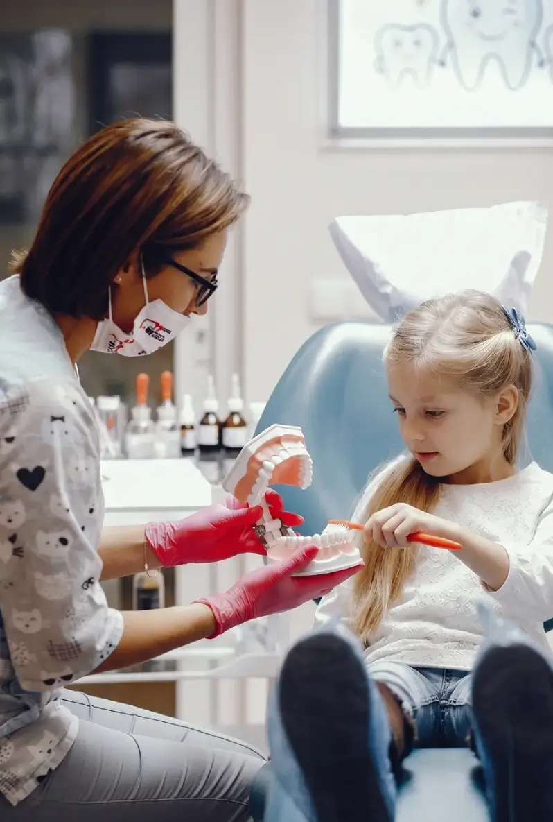 A dentist wearing a mask and gloves teaches a young girl to brush teeth using a model.