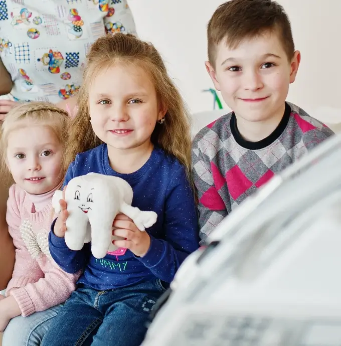 Three smiling children at a dentist's office, with one holding a tooth-shaped plush toy. The atmosphere is cheerful and comforting.