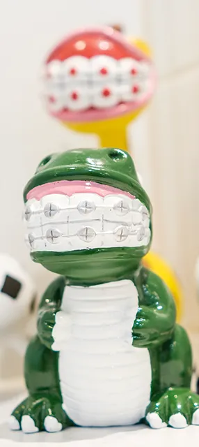 A green dinosaur figurine with a wide smile featuring braces on its teeth stands in front of a blurred yellow and red, similarly styled figurine. The scene is playful and whimsical.
