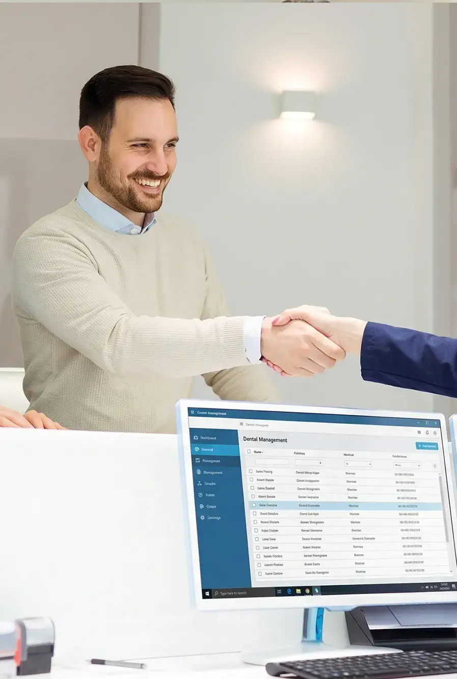 A man in a beige sweater smiles while shaking hands with someone in a blue suit. A computer screen in the foreground displays dental management system.