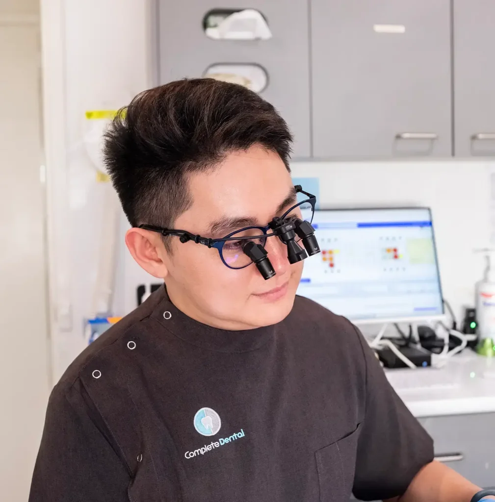 A dentist wearing magnifying loupes reviews patient information. Background shows a computer, dental tools, and cabinets, suggesting a modern clinic setting.