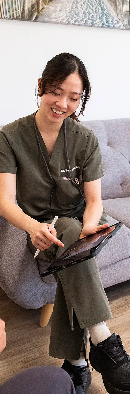 A woman in green scrubs sits on a gray sofa, smiling as she uses a stylus on a tablet. The setting is relaxed, suggesting a professional yet friendly atmosphere.