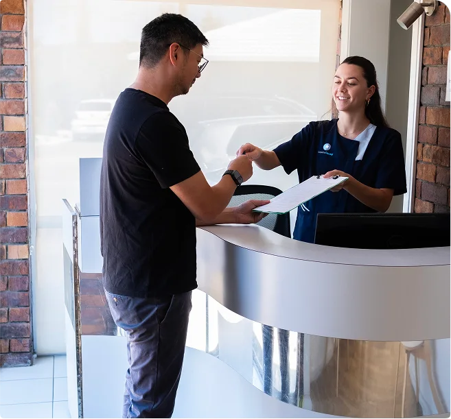 A receptionist hands a clipboard to a man in a modern office. The two are smiling